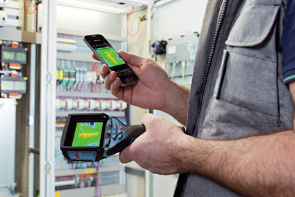 Person håller en smartphone och en termokamera framför ett öppet elskåp med kablar och komponenter.