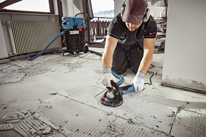 Person slipar golv med slipmaskin i rum, dammsugare i bakgrunden. Använder hörselskydd och handskar.