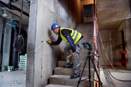 En person med blå hjälm och gul väst mäter en betongvägg med laser, omgiven av byggmaterial och verktyg.
