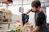 Man in workshop using a nail gun on wooden plank, wearing safety glasses and a black t-shirt. Shelves with boxes in the background.