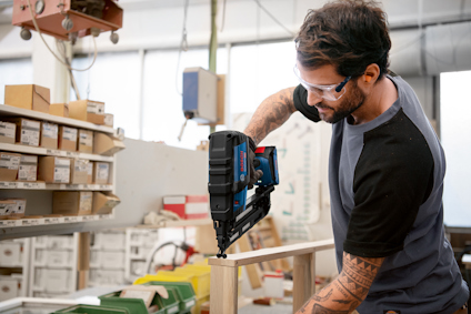 Man in workshop using a nail gun on wooden plank, wearing safety glasses and a black t-shirt. Shelves with boxes in the background.