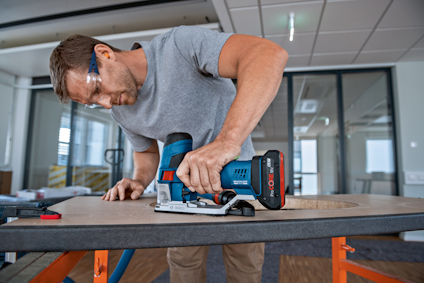 Person använder en blå sladdlös handöverfräs för att bearbeta en träyta inomhus.