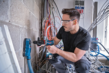 Man i skyddsglasögon borrar i betongvägg med en sladdlös borrmaskin, omgiven av kablar.