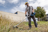 Person in protective gear using a grass trimmer on overgrown grass near a building with trees in the background.