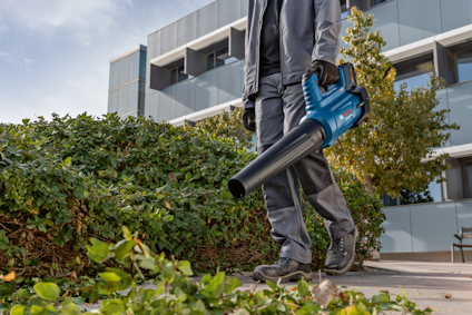 Person med lövblåsare går på trottoar bredvid buskar framför en modern byggnad.