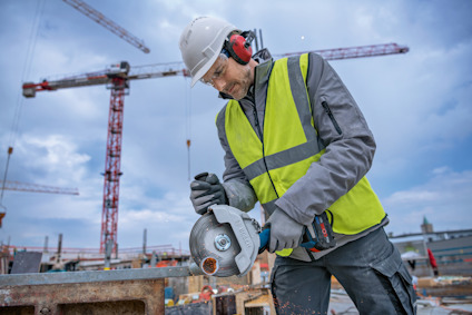 En person i skyddsutrustning använder en vinkelslip på en byggarbetsplats med kranar i bakgrunden.