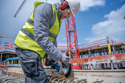 Person med hjälm och reflexväst sågar armeringsjärn på byggarbetsplats.