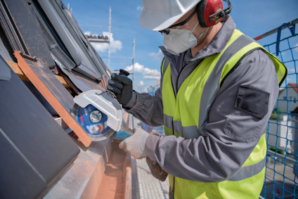 En person i varselväst använder en vinkelslip för att skära takpannor. Personen bär skyddshjälm och ansiktsmask.