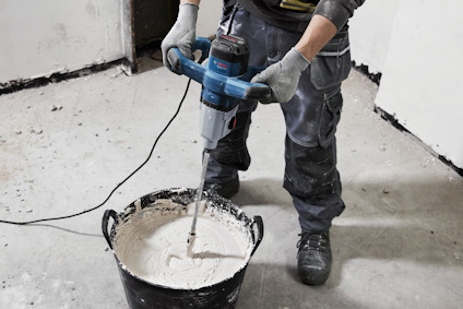 Person blandar vit vätska i hink med elmixer på ett golv, klädd i arbetsbyxor och handskar.