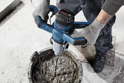 Person blandar cement med elektrisk mixer i grå tunnformad behållare. Handskar och arbetskläder synliga.