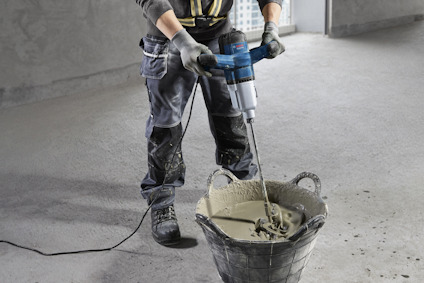 Person som använder en elektrisk borrmaskin för att blanda betong i en hink på ett betonggolv.