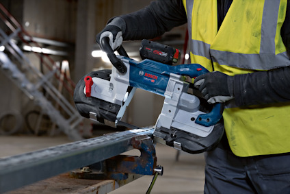 Person i gul väst använder batteridriven bandsåg för att skära genom metallstång på byggarbetsplats.