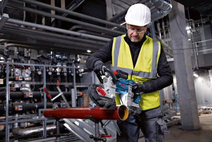 En person i gul skyddsväst och vit hjälm använder en kapmaskin på ett metallrör i en industriell miljö.