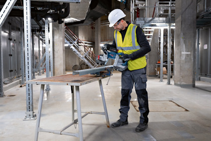 Person i skyddshjälm och väst, använder en elektrisk verktyg för att såga på ett stålbord i en industriell miljö.