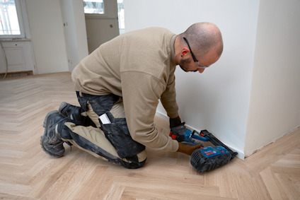 Person arbetar på parkettgolv, använder elverktyg vid väggen i ett rum.