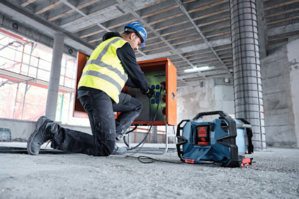 Person med skyddsutrustning ansluter kablar till orange skåp på byggarbetsplats, verktygsväska i förgrunden.