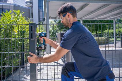 En person monterar en metallgrind med en trådlös borr, omgiven av grönska och byggnader i bakgrunden.