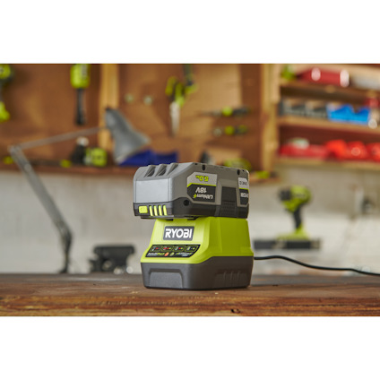En grön och svart Ryobi batteriladdare med batteri placerad på en arbetsbänk i verkstadsmiljö.