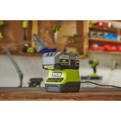 Ryobi batteriladdare och batteri på en arbetsbänk i en verkstad med suddiga verktyg i bakgrunden.