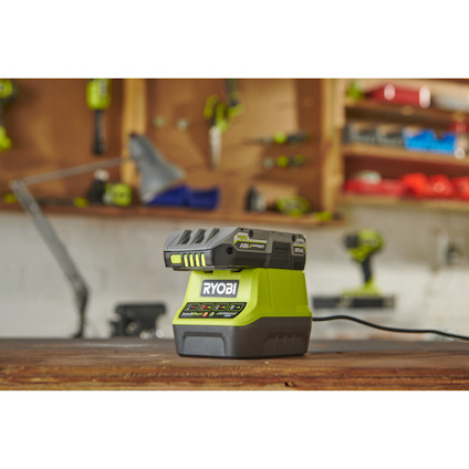 Ryobi batteriladdare och batteri på en arbetsbänk i en verkstad med suddiga verktyg i bakgrunden.