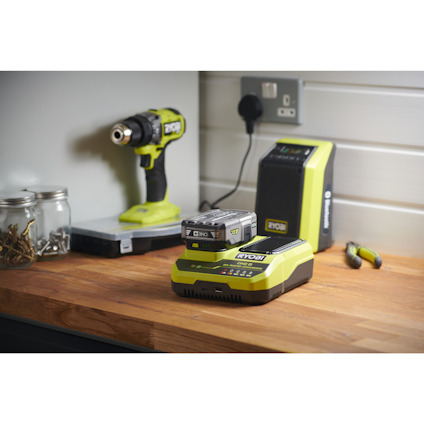 Snabbladdare av märket RYOBI med ett ONE+ batteri i på en träarbetsbänk. Elektriskt uttag, borrskryvdragare och högtalare i bakgrunden.