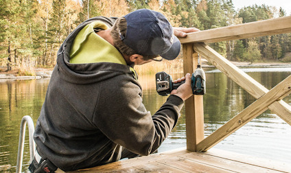Person skruvar på trästaket vid sjö, iklädd keps och munkjacka, omgiven av natur.