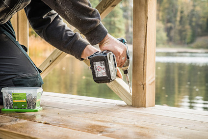 Person sätter ihop träkonstruktion vid sjö med en borrmaskin; skruvförpackning synlig.