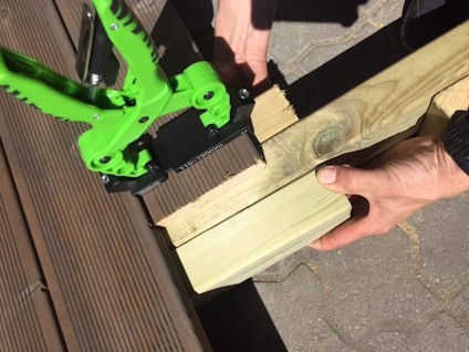 En person håller en träbit mot en regel och använder ett grönt verktyg för att placera en trallbräda utomhus.