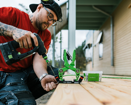 En person skruvar trall med en skruvdragare och använder ett grönt monteringsverktyg för att få rätt avstånd mellan brädorna.