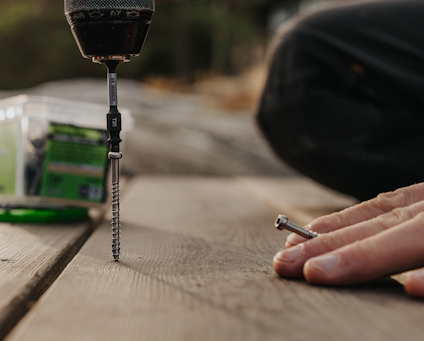 En person skruvar ner en skruv i en träplanka med en borrmaskin. En annan skruv hålls i handen bredvid.
