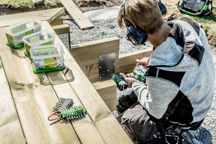 Person som arbetar på träkonstruktion med verktyg och skruvar i plastlådor. Bär hörselskydd.