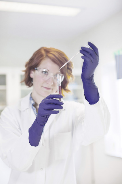 Person i laboratorium håller i ett provrör och en pipett, iklädd vit labbrock och blå handskar.