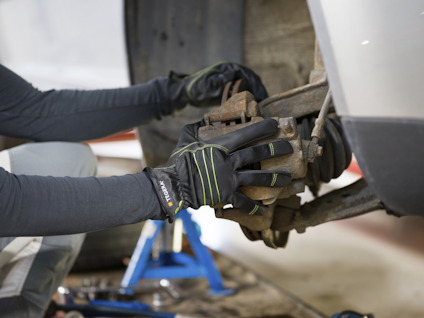 Person i arbetskläder och handskar arbetar med att reparera eller underhålla en bildel, bilen sitter upphöjd på en domkraft.
