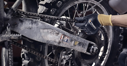 Närbild av en motorcykels kedja och bakhjul, där en person i handskar underhåller kedjan.