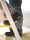 Person står på stege, bär svarta skyddsskor med märke "Jalas" och grå byxor i arbetsmiljö.