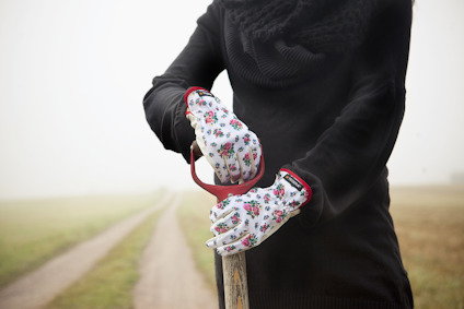 Person i svart tröja håller i en spade med röda handtag, klädd i blomstrade trädgårdshandskar, med landsväg i bakgrunden.