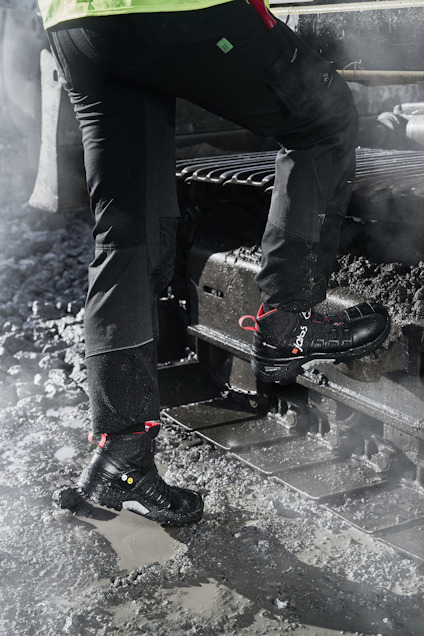 Person klädd i svarta arbetsbyxor och svarta skor med röd text, står på lerig trappa.