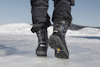 Svarta vinterstövlar med texten "Yalas" och dubbar på isigt underlag. Snö och berg i bakgrunden.