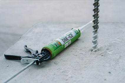 Patronspruta med en tub "ONE ICE" ankarmassa ligger på betong. Bredvid borrar en borr ett hål i betongen.