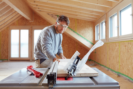 Man using table saw to cut wood planks in an attic under construction. Wearing safety glasses and casual clothing.