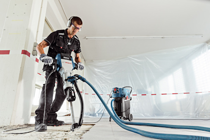 Person använder verktyg på en byggarbetsplats, omgiven av plastskynken och med skyddsutrustning.