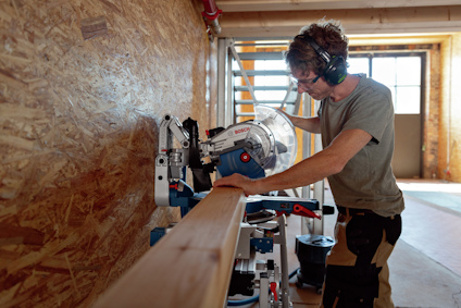 Person använder cirkelsåg för att såga träbräda i en verkstad, iklädd hörselskydd och skyddsglasögon.