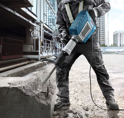 Person i arbetskläder använder en elektrisk bilningsmaskin på en betongyta. Byggnad i bakgrunden.