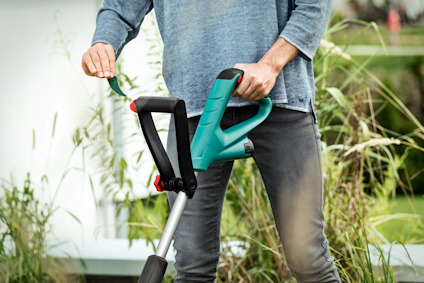 Person använder en elektrisk trimmer i trädgården, omgiven av högt gräs och några buskar.