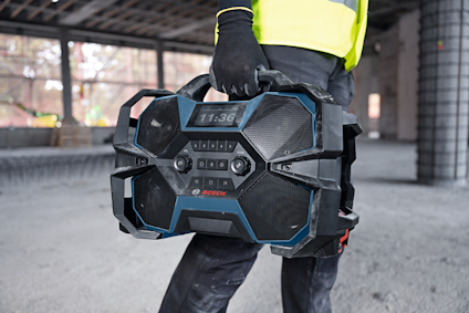 Person i arbetskläder håller en svart och blå bärbar musikspelare på en byggarbetsplats.