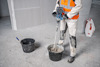 Arbetare i vit och orange skyddsutrustning blandar material i en hink med en mixer, betonggolv och väggskivor i bakgrunden.