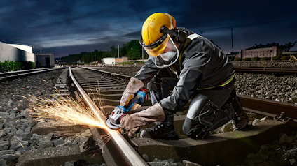 Person i skyddskläder och hjälm slipar järnvägsspår med en vinkelslip, gnistor flyger.