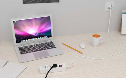 Bärbar dator på träbord med mus, penna, anteckningsblock, kaffekopp och förlängningssladd ansluten i väggen.