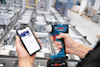 Person håller en Bosch skruvdragare och en smartphone med en app på en fabriksgolvmiljö.