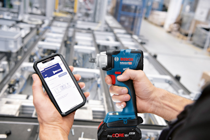Person håller en Bosch skruvdragare och en smartphone med en app på en fabriksgolvmiljö.